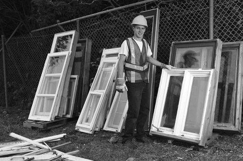 A male worker in a safety helmet and reflective vest stands outdoors on a grassy area near a chain-link fence, holding a large white-framed window with glass panels. Behind him, several other windows with white frames and glass panes are stacked upright on the ground and leaning against the fence, indicating accumulated or removed property items. The windows vary in size, and some appear to be new or in good condition, with clean, smooth finishes and clear glass sheets. The setting suggests a site involved in rubbish removal or disposal of building waste, typical of properties in Belmont or the surrounding postcode area, with the environment illuminated by natural daylight. The scene is professional and neutral, representing typical waste removal activities related to property renovation or clearance, handled by companies like Commercial Waste Removal Belmont specializing in rubbish disposal services.
