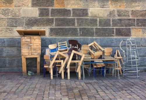 A collection of discarded wooden furniture, including small chairs, tables, and a wooden cabinet, arranged outdoors against a grey brick wall. The furniture appears to be a mix of unfinished and weathered wood, with some pieces leaning or stacked haphazardly. To the right, there is a metal shelving unit with a simple, open design. The ground is paved with cobblestones, and the overall scene suggests a setting where old or unwanted furniture has been left for rubbish removal. The image is relevant for a waste management service operating in Belmont, familiar with handling various types of furniture waste from residential or commercial properties.