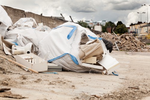 A large pile of mixed construction waste and debris, including white polystyrene panels, plastic piping, and cardboard packaging, scattered on a construction site. The waste is contained within a blue and white bag and surrounding materials are piled on a dirt surface, with a background of an unfinished wall, loose rubble, and distant buildings under a cloudy sky. The scene illustrates typical rubbish accumulated during building or renovation projects, with the rubble and debris positioned on level ground in an outdoor environment consistent with a construction or demolition area in the vicinity of Belmont. The presence of construction elements like piping and packaging materials highlights waste common in commercial or residential renovation settings, aligning with rubbish removal services offered by Commercial Waste Removal Belmont in the local area.