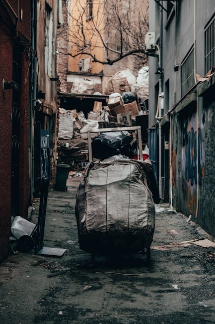 A waste collection vehicle parked on a narrow urban street adjacent to old stone buildings with multiple stories. The truck is white with signs of rust and grime on its rear compartment, which is open