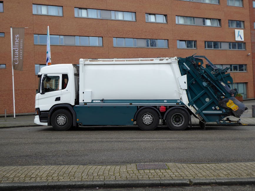 A large blue wheeled recycling bin made of plastic with a textured surface and a closed lid, positioned on a concrete pavement in front of a white delivery van or storage vehicle. The bin is situated 