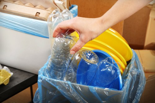 A close-up of a person's hand holding a clear plastic water bottle with a partially crushed body, positioned over a large blue plastic bin lined with a transparent rubbish bag. Inside the bin are several yellow and blue plastic buckets stacked together, with the yellow bucket visibly atop the blue bucket. The background shows a wooden surface and a piece of furniture, indicating this scene takes place indoors or in an enclosed space suitable for waste collection. The image captures the act of sorting or preparing waste materials, relevant to commercial rubbish removal services such as those provided by Commercial Waste Removal Belmont in the Belmont area. The textures of the plastic items are smooth and shiny, with the discarded bottle appearing crinkled, emphasizing the nature of waste materials. The lighting is neutral and even, highlighting the objects clearly without shadows or glare, underpinning a professional and factual visual presentation aligned with waste management practices in the UK.